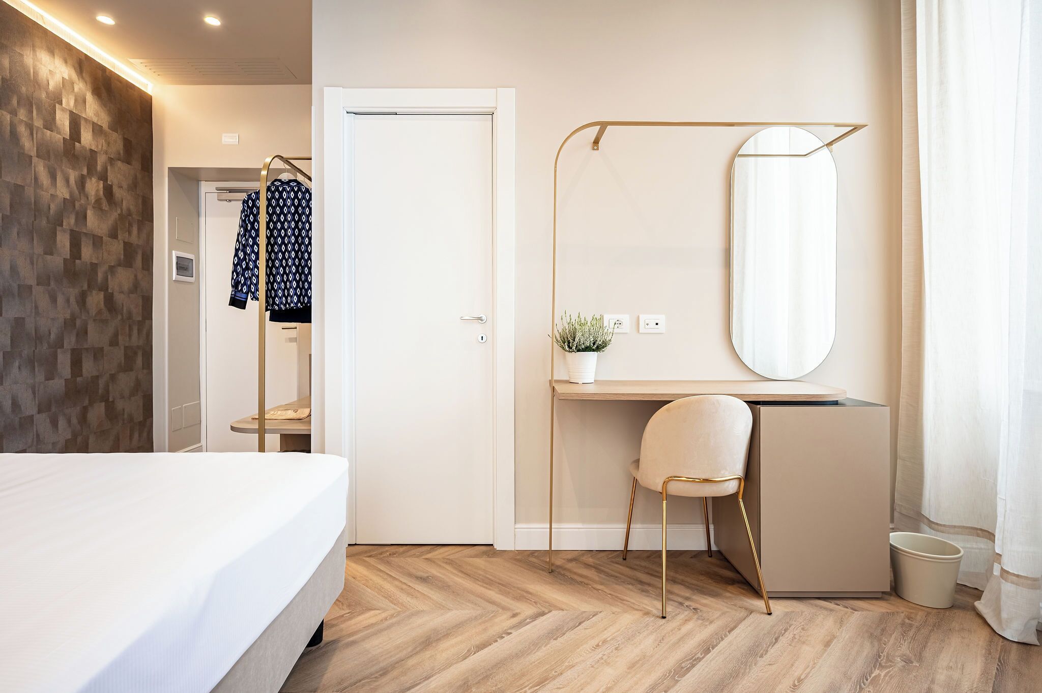 Una habitación de hotel moderna con cama, escritorio con silla, un espejo grande y una pequeña planta en maceta.