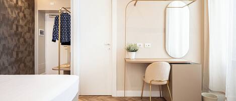 A modern hotel room with a bed, a desk with a chair, a large mirror, and a small potted plant.
