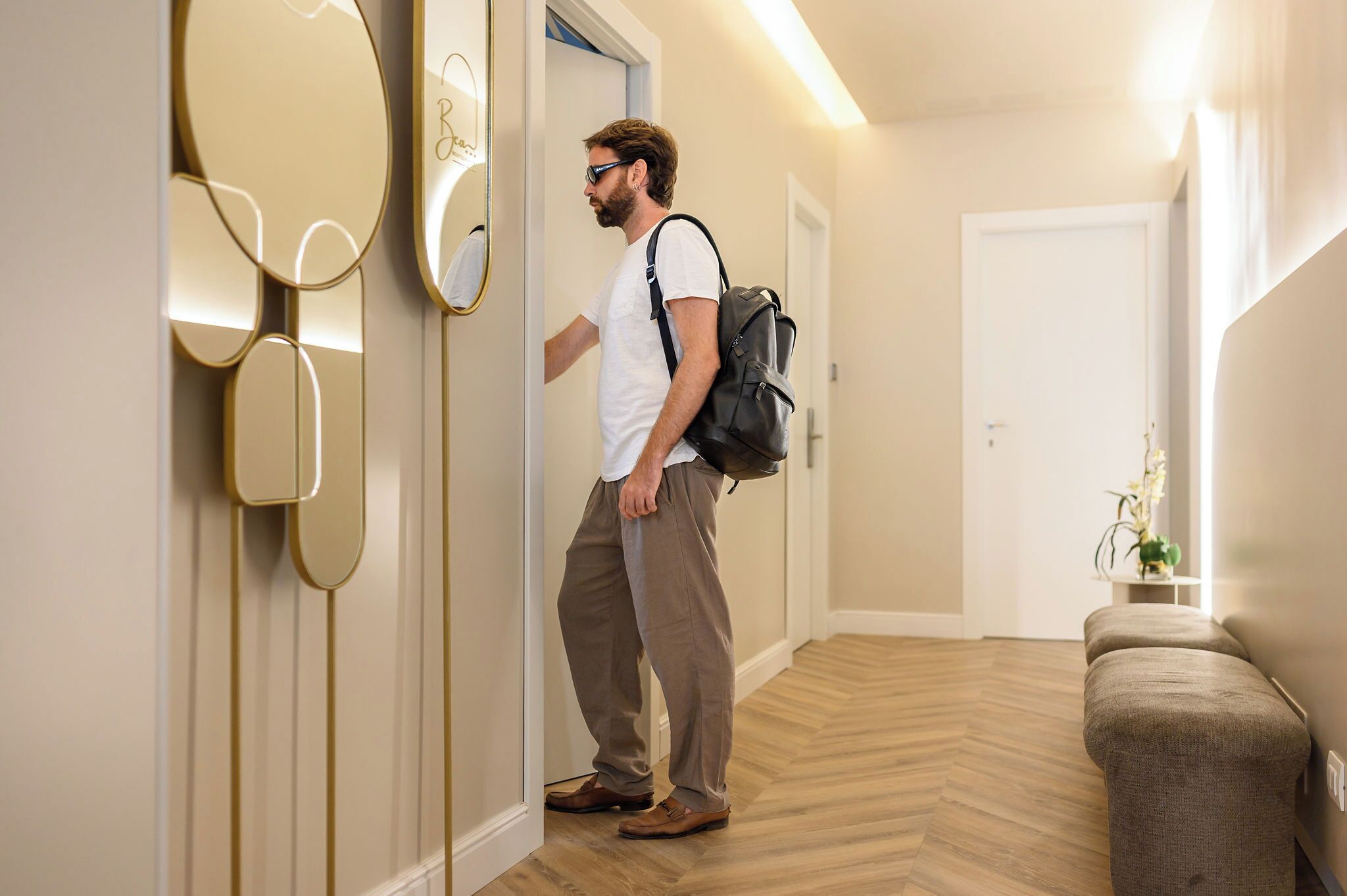 A man in a hallway with a mirror, a bench, and a door.