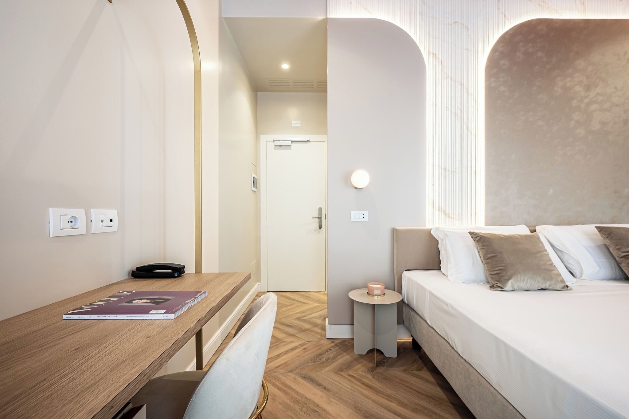A modern hotel room with a wooden desk, a bed with beige and white bedding, a small round table with a candle, and a door leading to another room.