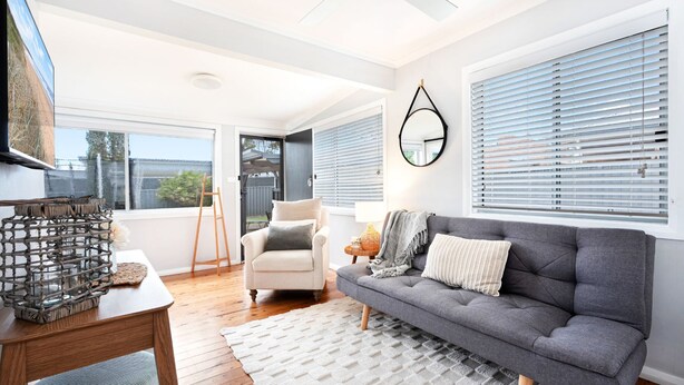 A modern living room with a grey sofa, a white armchair, and a wooden coffee table. There is a large window with blinds, a hanging light fixture, and a view of the outdoors.