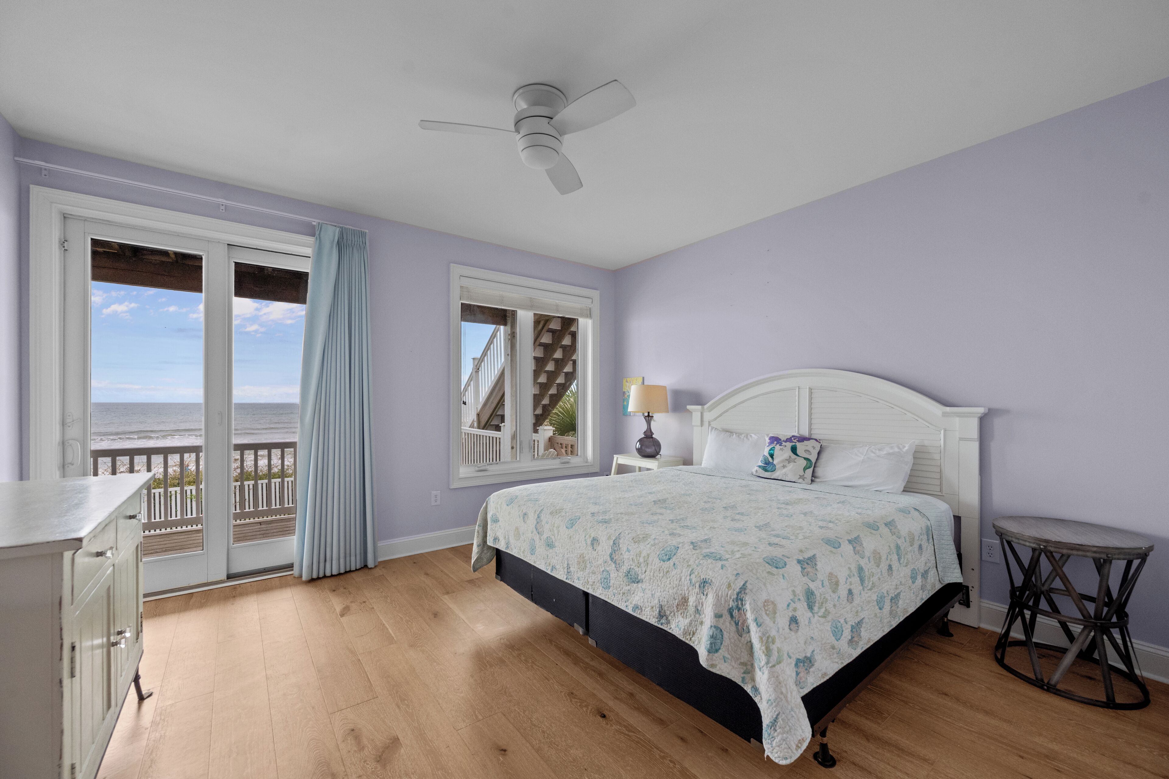 A bedroom with a bed, a desk, a chair, a ceiling fan, and a view of the ocean through a sliding door.