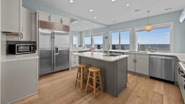 A modern kitchen with stainless steel appliances, wooden stools, and large windows overlooking a scenic view. - North Shore Paradise #199 - 7 BR, 7.5 BA Oceanfront Home w/ Pool & Hot Tub (Surf City)