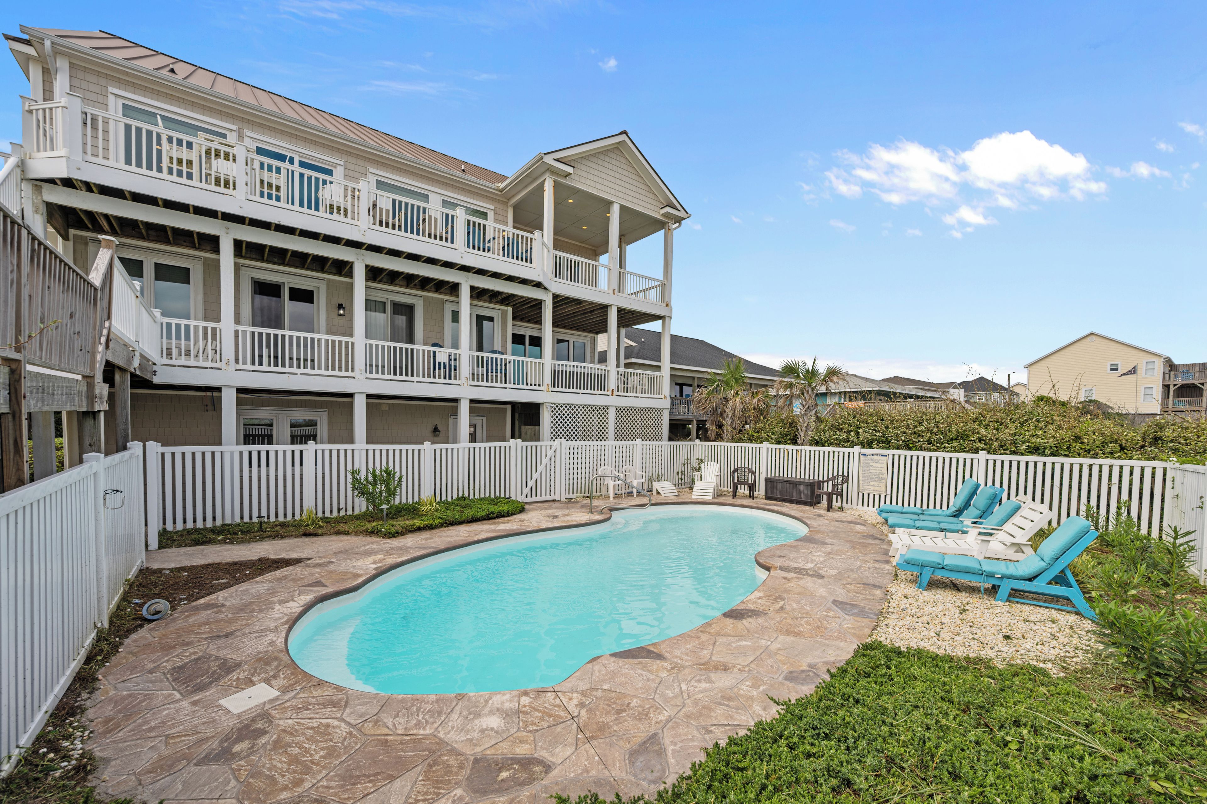 A two-story house with a swimming pool, a white fence, and a patio area with lounge chairs.