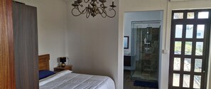 Ein Schlafzimmer mit Bett, Nachttisch, Lampe und einer Chandelier. Eine Glastür führt zu einem Duschbereich mit Glastür und Blick ins Freie.