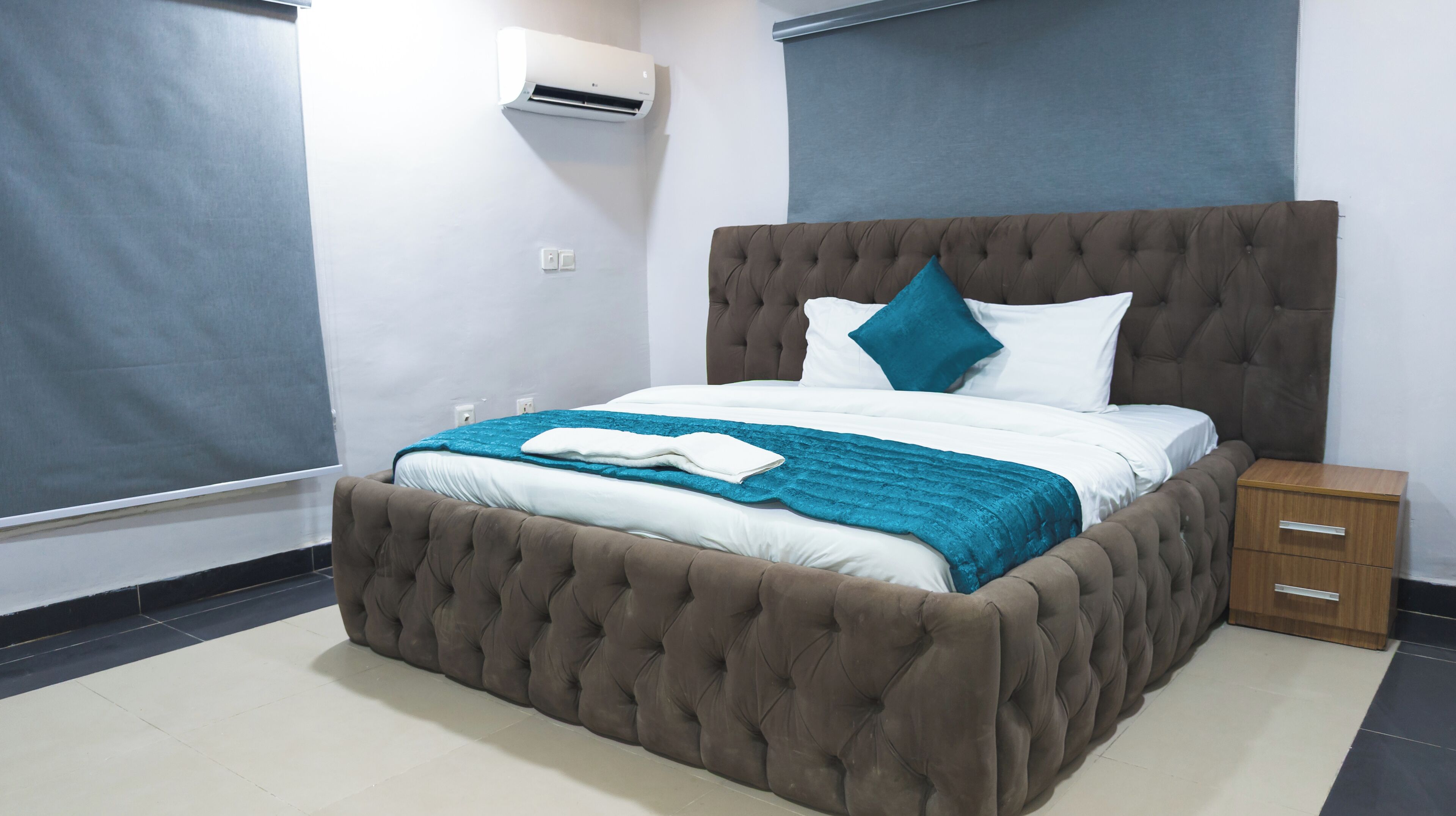A bedroom with a tufted headboard, a bed with white and teal bedding, a wooden nightstand, and a wall-mounted air conditioner.