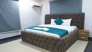 A bedroom with a tufted headboard, a bed with white and teal bedding, a wooden nightstand, and a wall-mounted air conditioner.
