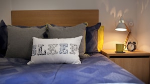 A neatly made bed with a pillow featuring a bold design, a bedside table with a lamp and an alarm clock, and a wall socket. - Student Only Algernon Firth (Leeds)