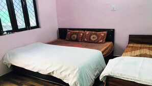 A room with two beds, one with a white bedspread and the other with a patterned bedspread. The room has a wooden floor and a window with a metal grill. - Hotel Rama Palace (Ukhimath)
