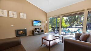 Un salon avec une cheminée, un téléviseur et un canapé. Une grande fenêtre offre une vue sur les arbres et donne sur un patio à l’extérieur.