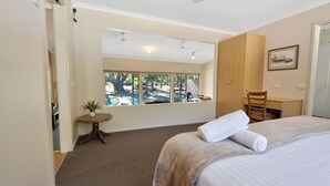 Une chambre d’hôtel avec un lit, un bureau, une chaise, une petite table avec une plante et une fenêtre donnant sur des arbres.