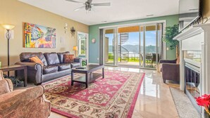 Uma sala de estar ampla com um sofá, mesa de centro e um tapete grande. Há um ventilador de teto, um quadro na parede e vista para o exterior através de uma porta de correr de vidro.