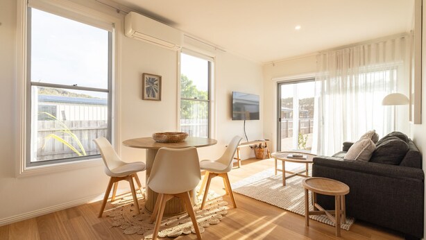 Ein modernes Wohnzimmer mit einem runden Esstisch, weißen Stühlen, einem dunklen Sofa und einem an der Wand befestigten Fernseher.