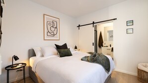 Un dormitorio moderno con una cama grande, una mesita de noche con lámpara, un cuadro abstracto enmarcado y una puerta corredera que da a un baño.