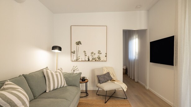 Una sala de estar con un sofá, una silla, una lámpara de pie, un cuadro enmarcado y un televisor montado en la pared.