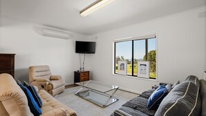 Una sala de estar con un sofá color beige, un sofá gris oscuro, una mesa de centro de vidrio, un sillón color beige, un televisor montado en la pared y un amplio ventanal con vistas a vegetación y un edificio.