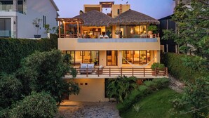 A two-story house with a thatched roof, a balcony, and a well-maintained garden. - 3BR Luxury Villa | Pool & Panoramic Ocean Views (Huatulco)
