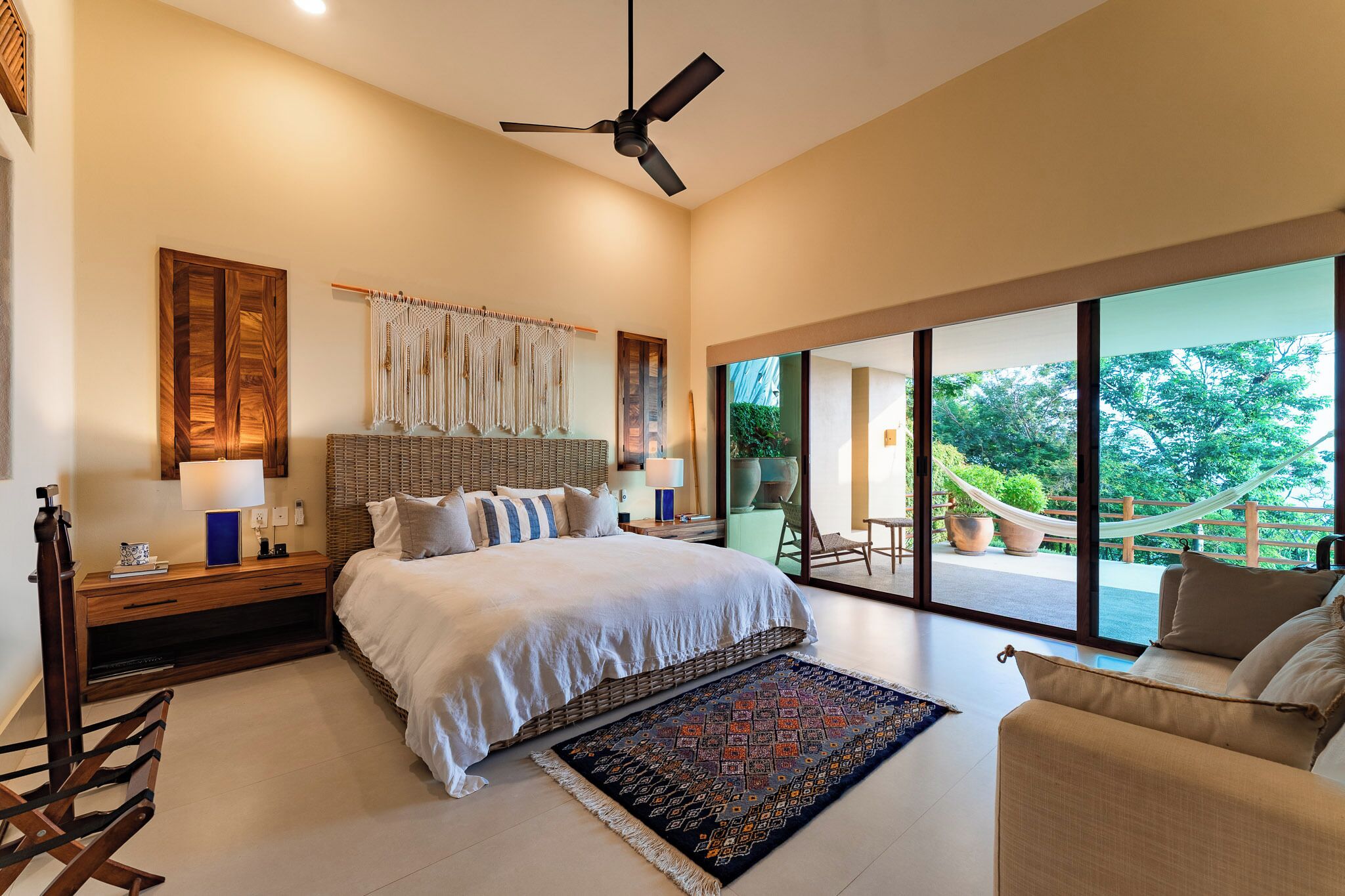 Une chambre spacieuse avec un grand lit, un ventilateur de plafond, une table de chevet, une commode, une fenêtre donnant sur un paysage verdoyant et un balcon avec un hamac.