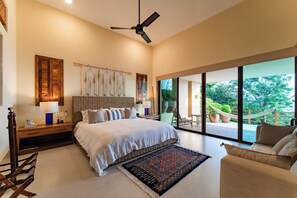 A spacious bedroom with a large bed, a ceiling fan, a nightstand, a bedside table, a window with a view of greenery, and a balcony with a hammock.