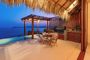 A covered outdoor area with a pool, wooden furniture, and a view of the sea.