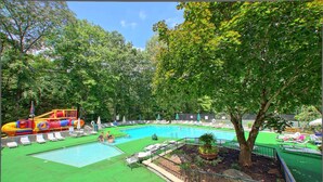 Una piscina con tobogán y zona de juegos, rodeada de árboles y sillones de descanso.