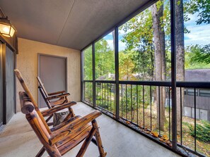 A wooden bench with a metal railing on a balcony with large windows overlooking a wooded area.