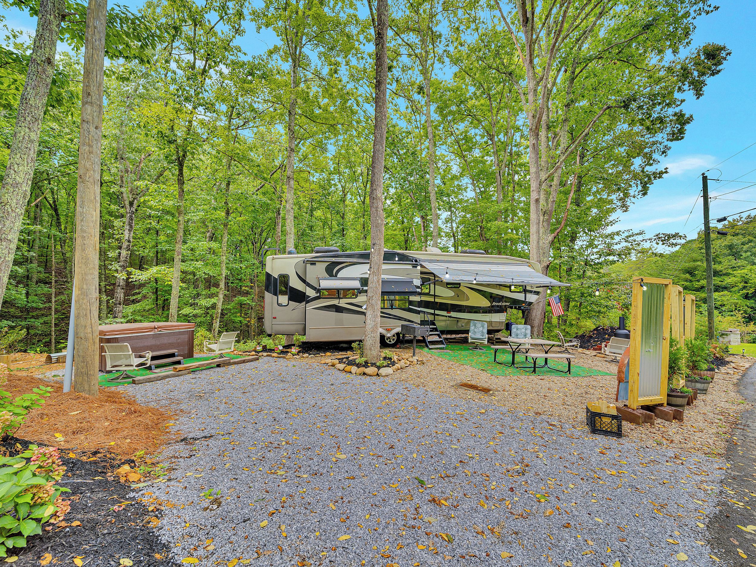A recreational vehicle (RV) parked in a forested area with a gravel path, surrounded by trees and a picnic table.