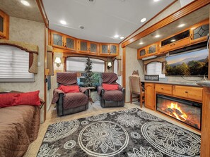 A cozy RV interior with two cushioned chairs, a small Christmas tree, a fireplace, and a television displaying a scenic view.
