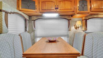 A compact RV interior with a dining table, cushioned seating, wooden cabinets, and a window with blinds.