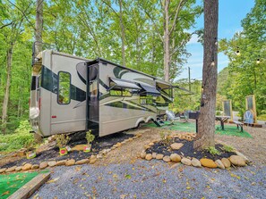 A recreational vehicle parked in a forested area with a gravel path, surrounded by trees and outdoor furniture.