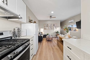Una cocina moderna con estufa, refrigerador y armarios blancos. Hay un comedor con mesa y sillas, y una sala de estar con sofá y una mesita de centro.