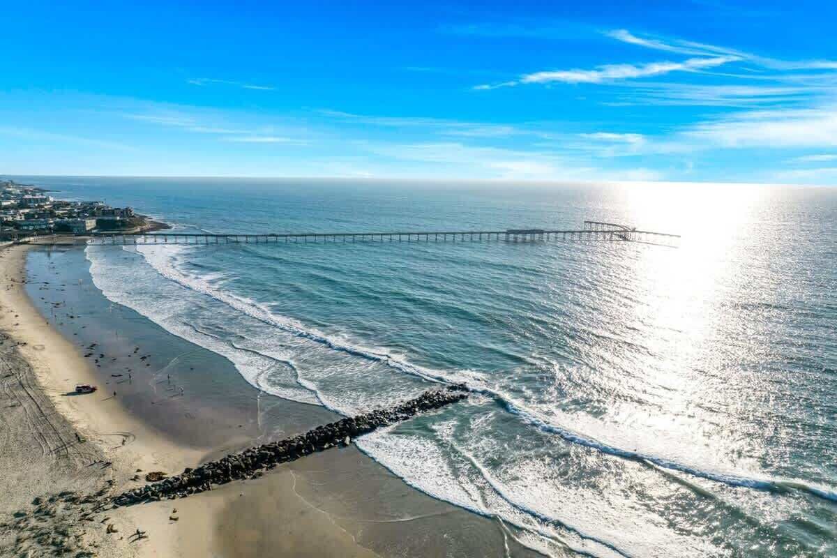 Ein Küstenstrand mit einem Steg, der sich ins Meer erstreckt, sandigem Ufer und blauem Himmel.