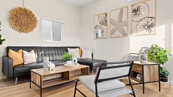 Ein modernes Wohnzimmer mit einem schwarzen Ledersofa, einem Holztisch, einem Stuhl mit Metallgestell und einem Beistelltisch aus Holz. An der Wand befinden sich gerahmte Pflanzenbilder und ein gewebter Wandbehang.