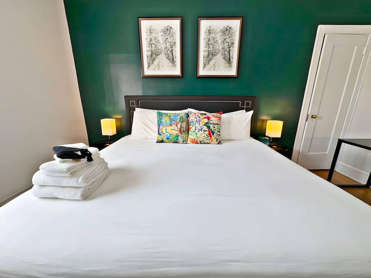 A hotel room with a neatly made bed, two framed artworks on the wall, and a stack of towels on the side.