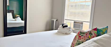 A hotel room with a bed, a radiator, a window with blinds, and a stack of folded towels.