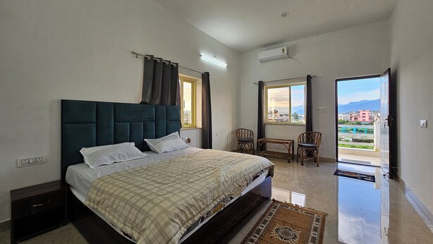 Quarto com cama, duas cadeiras, uma mesa pequena, janela com cortinas e vista para edifícios e montanhas.