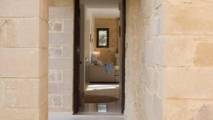 A stone-walled hallway leading to a room with a bed, a small table, and a window.