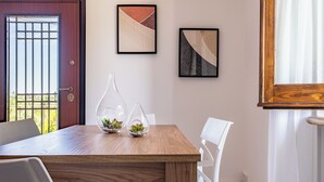 Un comedor con una mesa de madera, sillas blancas y dos jarrones de vidrio con plantas. Se observan dos obras de arte enmarcadas en la pared y una puerta con reja de metal.