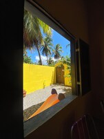 Vista pela janela: uma parede amarela, uma porta e uma palmeira.