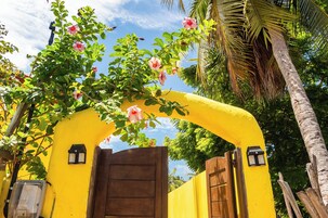 Um arco amarelo com porta de madeira, rodeado por vegetação exuberante e flores cor-de-rosa.