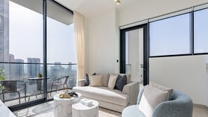 Un salon moderne comprenant un canapé, un fauteuil et une table basse, de grandes fenêtres offrant une vue sur la ville, ainsi qu’un balcon avec des sièges d’extérieur.