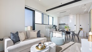Un salon moderne avec un canapé, un pouf et une table basse. On y trouve également un coin repas avec une table et des chaises. La cuisine est visible au fond, avec des meubles blancs et une grande fenêtre.