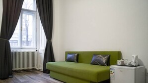 Een kamer met een groene bank, een kleine witte koelkast, een raam met gordijnen en een radiator.