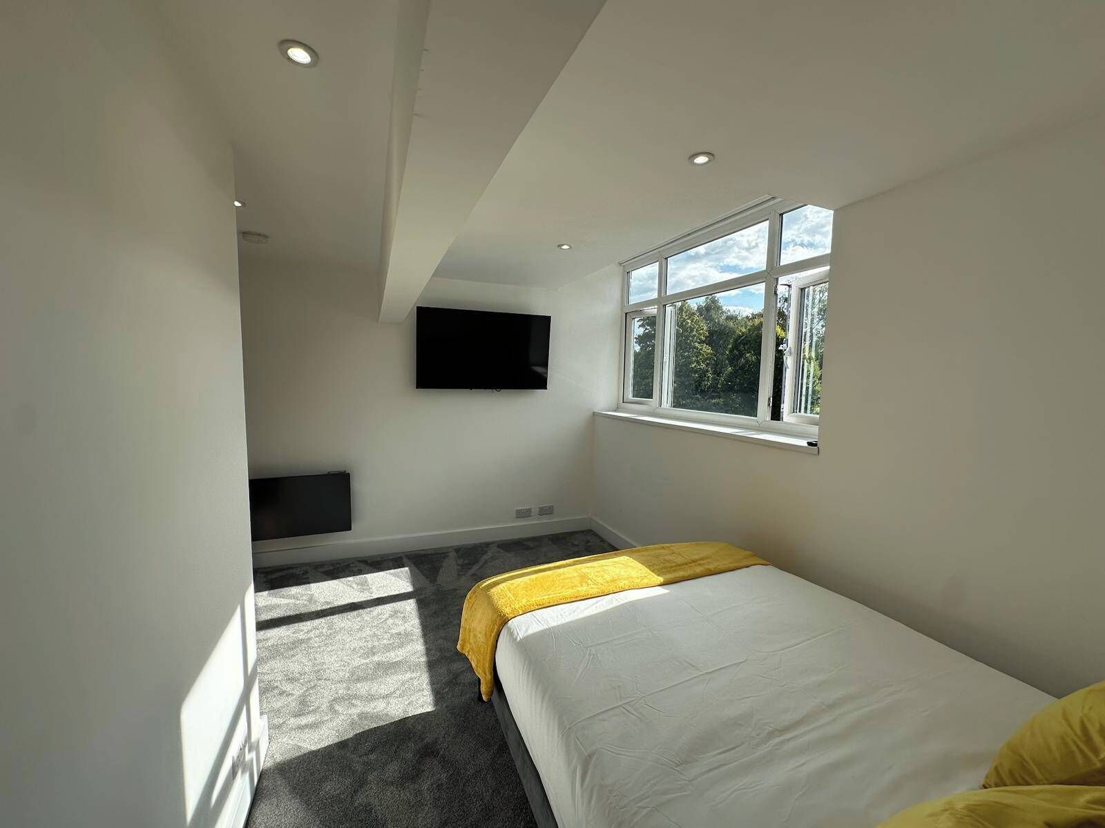 A bedroom with a bed, a TV mounted on the wall, a window, and a carpeted floor.