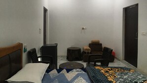 A small, dimly lit room with a bed, a desk, a chair, a sofa, and a patterned rug. There is a door labeled "WASH ROOM" and a small table with a stool. - Wilder Lust Homestay (Bhira)