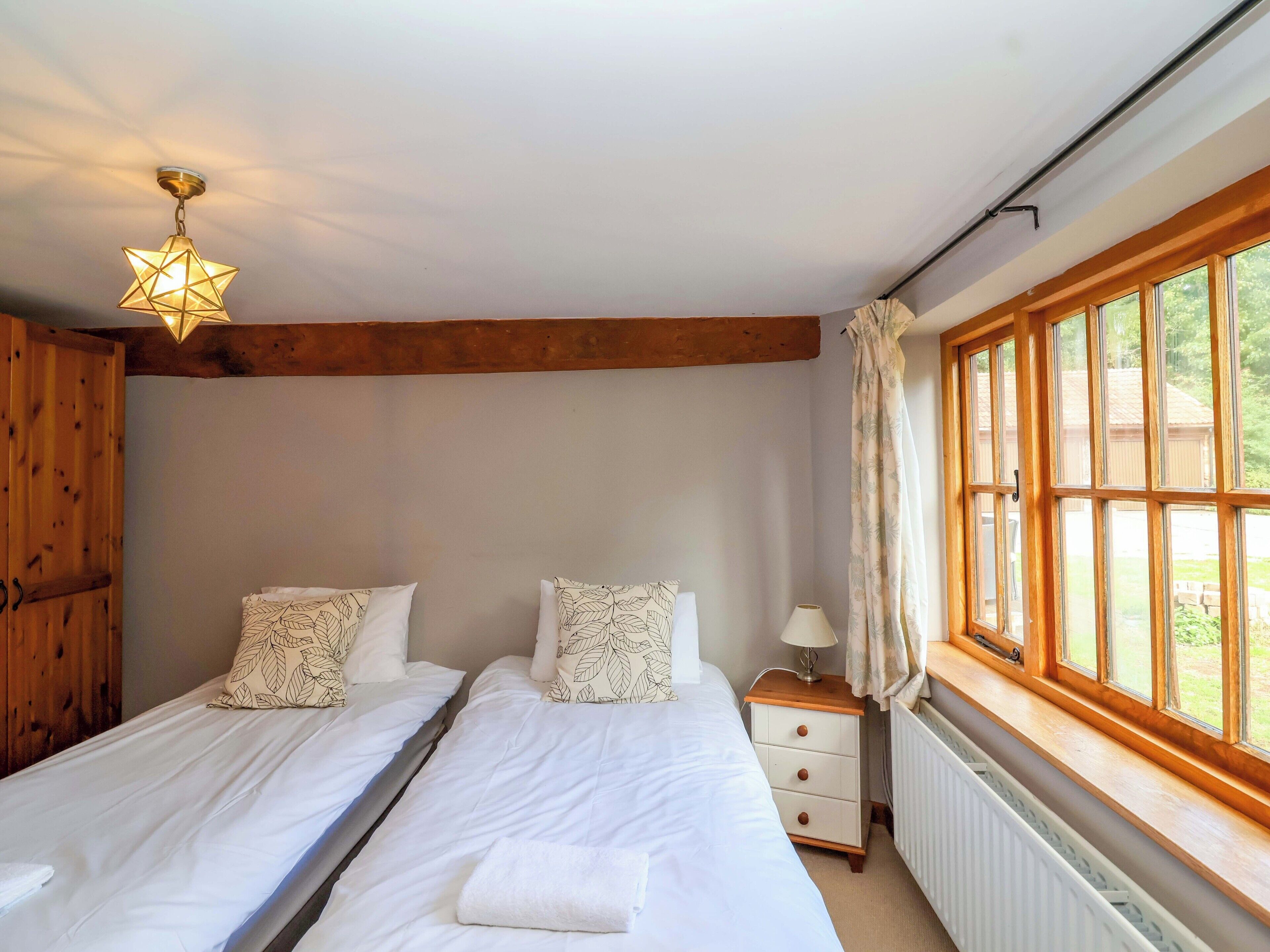 A room with two single beds, a nightstand with a lamp, a wooden ceiling beam, and a window with curtains.