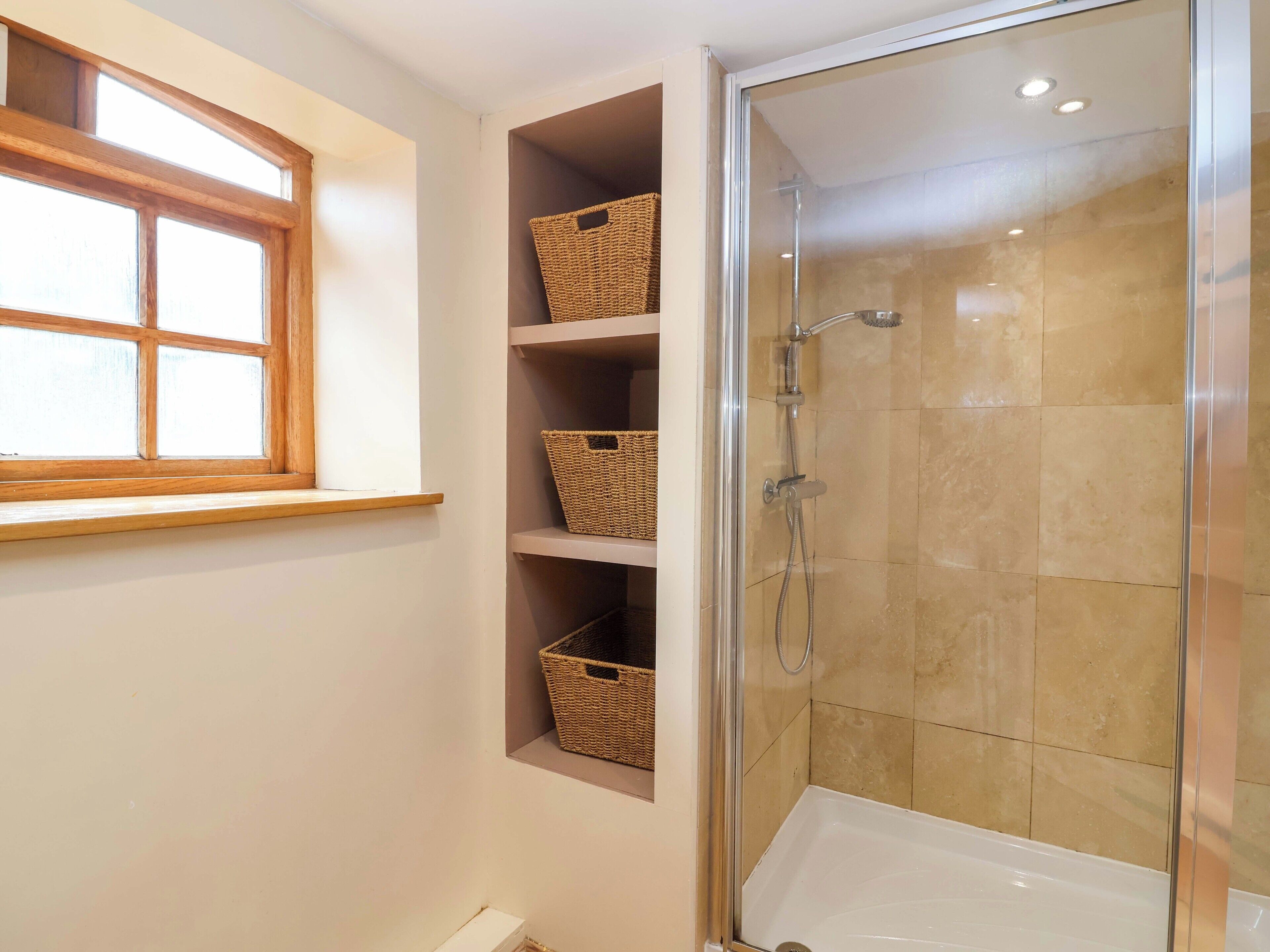 A bathroom with a glass-enclosed shower, a window with a wooden frame, and shelves with woven baskets.