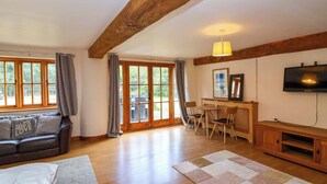 A cozy living room with a wooden ceiling, a sofa with a cushion, a TV, and a dining area with chairs. - Millbarn (Taunton)
