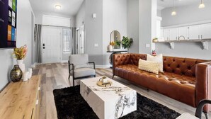 Un salon moderne avec un canapé en cuir marron, une table basse blanche et un tapis noir. On y trouve une porte blanche et un coin cuisine avec des meubles blancs.