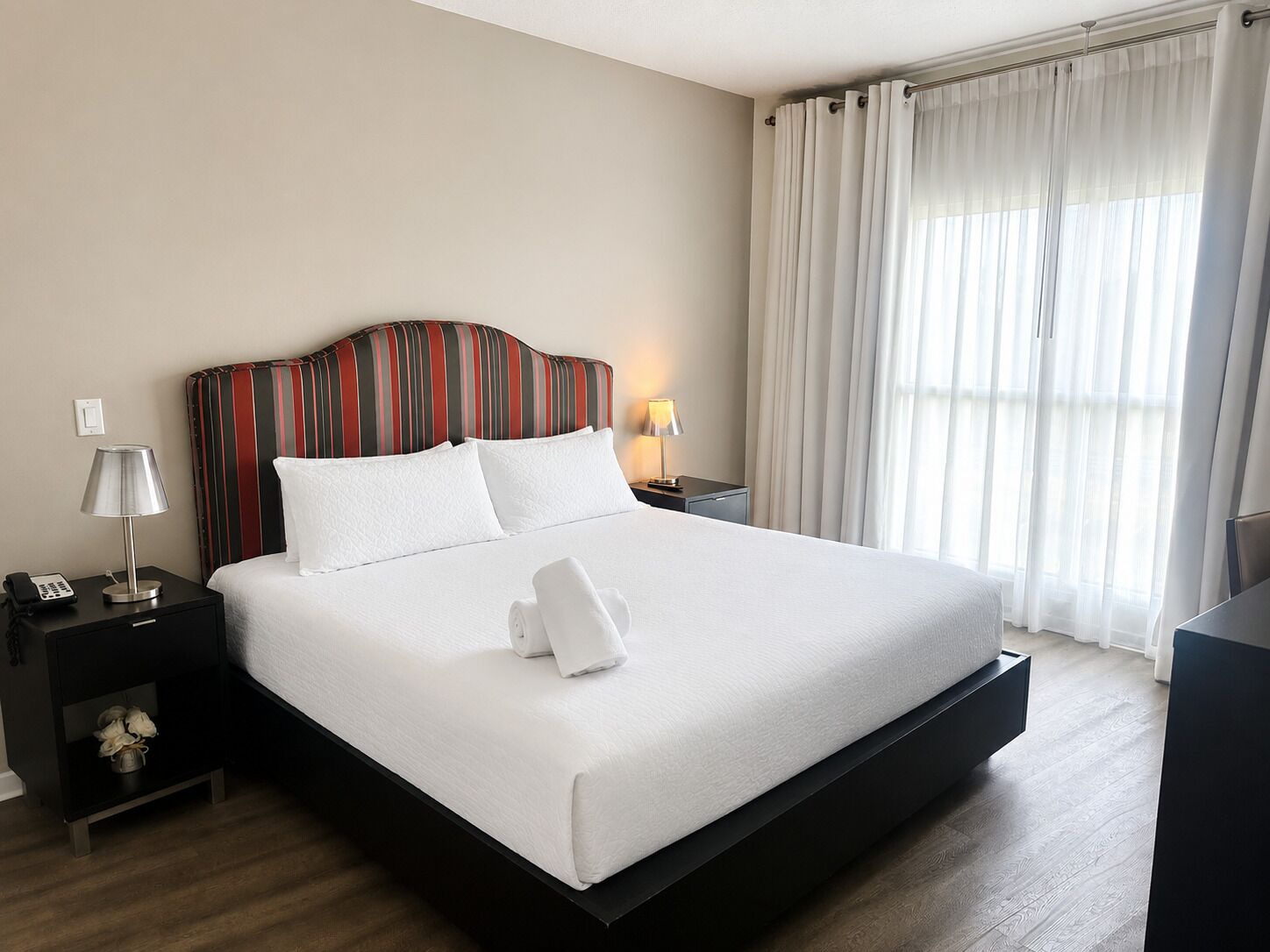Une chambre d’hôtel avec un grand lit, une tête de lit à rayures, une lampe de chevet, une table de chevet et une fenêtre avec des rideaux.